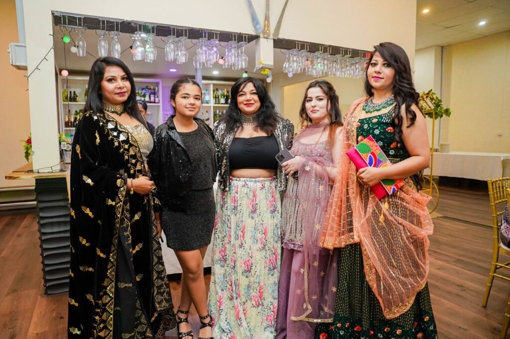 Home Group of women in colorful attire at a festive indoor party, smiling and posing for a photo.